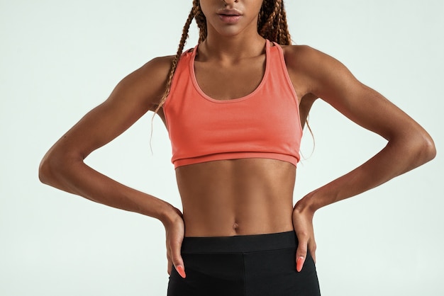 Perfect body. Cropped photo of muscular young african woman in sportswear keeping arms on hips while standing against grey background. Sport concept. Human body