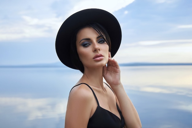 Perfect brunette beauty woman in a black hat and a black dress poses near a lake against a blue sky. Long hair woman and beautiful beauty makeup on her face