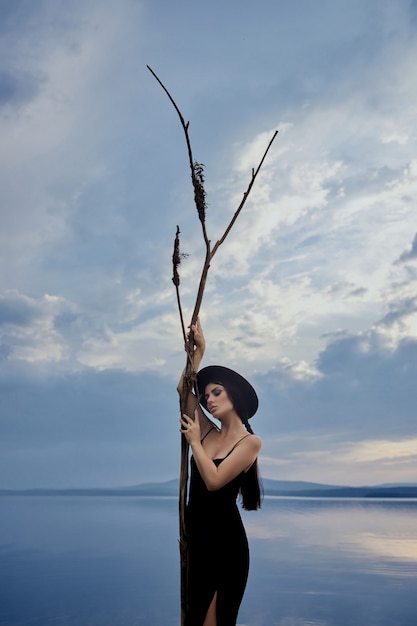 Perfect brunette beauty woman in a black hat and a black dress poses near a lake against a blue sky.