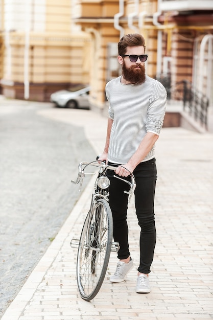 Perfect day for a ride. Full length of young bearded man rolling his bicycle and looking away while walking outdoors