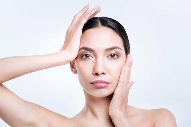 Perfect gezicht. Mooie donkerharige vrouw raakt haar wang aan met een hand en raakt haar hoofd aan met een andere terwijl ze tegen een witte muur poseert