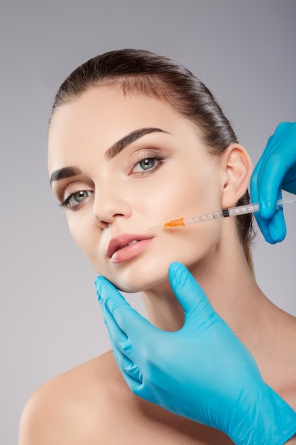 Perfect girl with thick eyebrows at studio background, doctor's hands wearing blue gloves near patient's face, holding syringe near face, looking at camera.