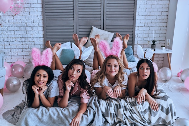 Идеальные девушки. Вид сверху четырех игривых молодых женщин в кроличьих ушах, корча гримасу и улыбающихся, лежа на кровати