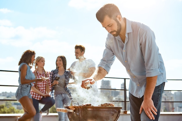 完璧な肉バーベキュー若いひげを生やした男が立っている間グリルで肉をバーベキュー