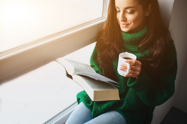 Perfect morning a young brunette woman sits on a windowsill and\
holding a book and sa cup of tea in her hands. female model dressed\
in green oversized sweater.