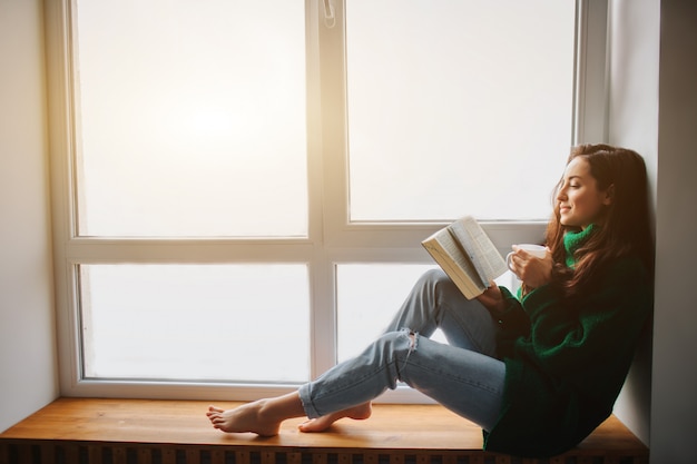 Mattina perfetta una giovane donna bruna si siede su un davanzale e tiene in mano un libro e una tazza di tè. modello femminile vestito in maglione oversize verde.