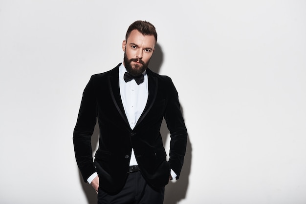 Perfect style. Handsome young man in full suit keeping hands in pockets and looking at camera while standing against grey background