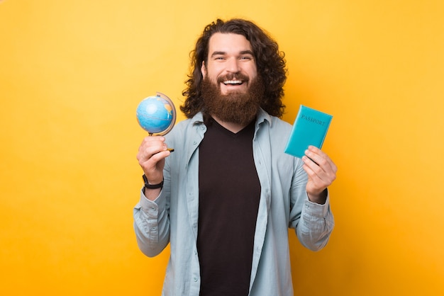 Perfect time to travel the world, cheerful bearded hipster man holding globe and passport over yellow background
