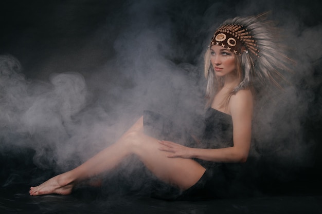 Perfect woman in garb of American Indians in smoke on a gray background. A hat made of feathers. Mysterious mystical way, sexy body, beautiful back. Attractive blonde with a beautiful face