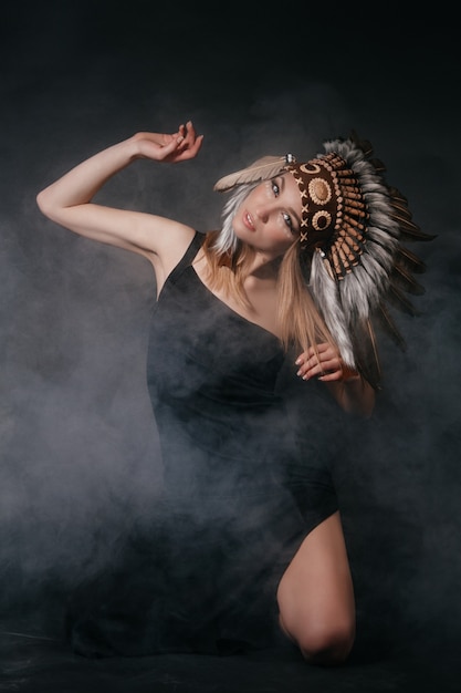 Идеальная женщина в одежде американских индейцев в дыму на сером фоне. Шапка из перьев. Загадочный мистический образ, сексуальное тело, красивая спина. Привлекательная блондинка с красивым лицом