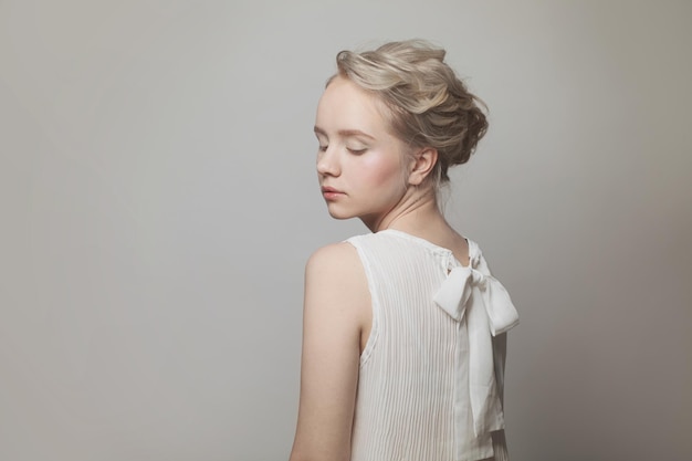 Perfect young blonde woman with gentle updo hair in white bow back blouse on white background