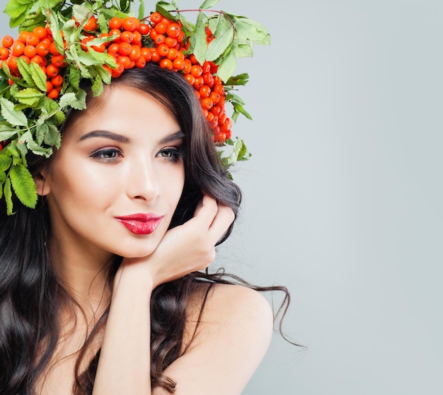 Perfect young brunette woman with long wavy hair makeup and red berries and green leaves Beautiful female face