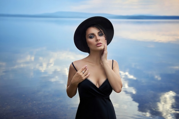 Perfecte brunette schoonheidsvrouw in een zwarte hoed en een zwarte jurk poseert in de buurt van een meer tegen een blauwe hemel. Lange haarvrouw en mooie schoonheidsmake-up op haar gezicht