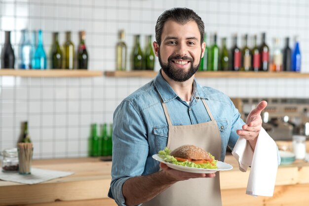 Perfecte lunch. Knappe bebaarde positieve ober die gelukkig kijkt terwijl hij aan het werk is en met een lekkere grote sandwich op een bord in zijn hand staat