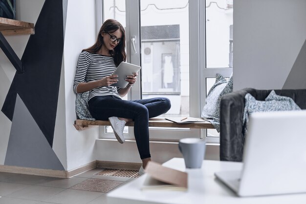 Perfecte plek om te werken. Aantrekkelijke jonge vrouw die digitale tablet gebruikt terwijl ze op de vensterbank zit