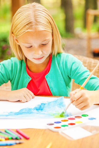 Performing her creativity. Cute little girl drawing something on paper while sitting at the table and outdoors