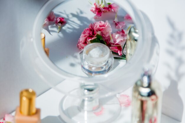 Perfume bottle, skin care cream and dianthus flowers around on white wall