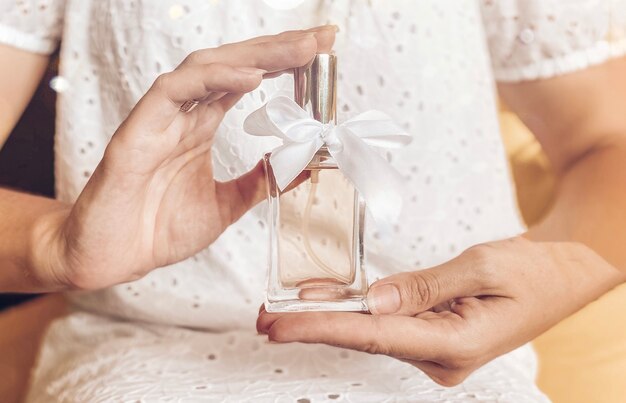 Perfume bottle with a white gift ribbon in a female hand against the background of a girl in a white dress in pastel light colors.Eau de toilette, eau de parfum, beauty concept.