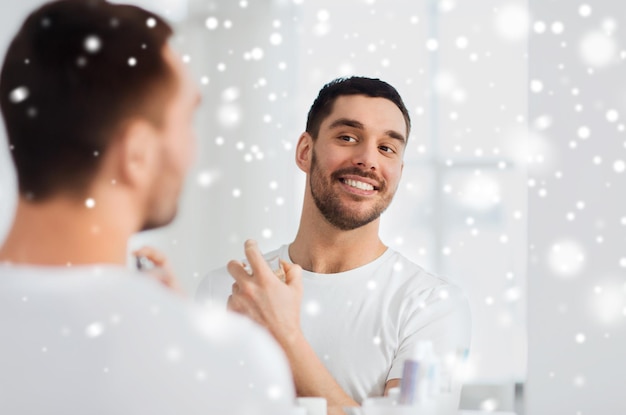 Photo perfumery, beauty and people concept - happy smiling young man with perfume looking to mirror using scent at bathroom over snow