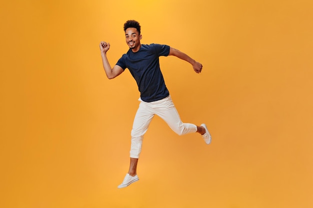 Perky man in light pants jumping and having fun on orange background Snapshot of brunette guy in white trousers posing on isolated