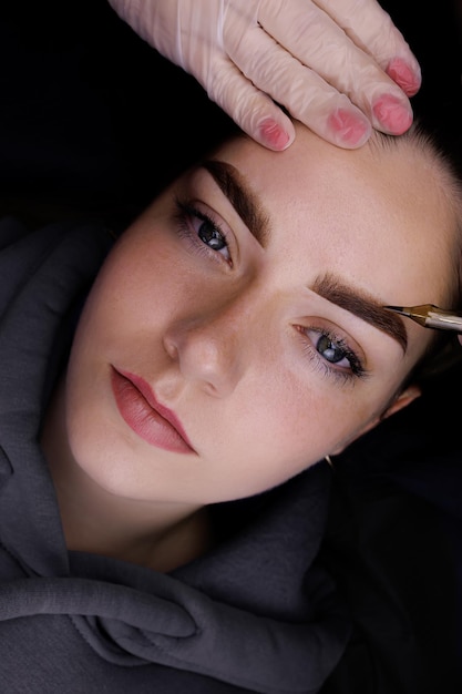 Permanent eyebrow makeup the master holds a tattoo machine near the model's eyebrows showing the result after tattooing