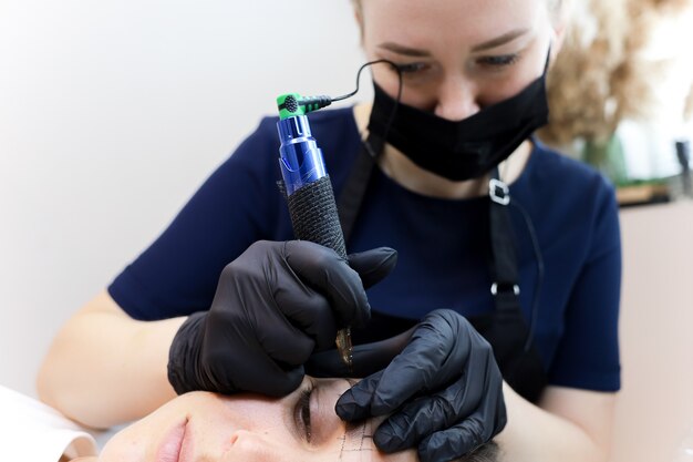 The permanent makeup artist leans over the client and performs an eyebrow tattoo