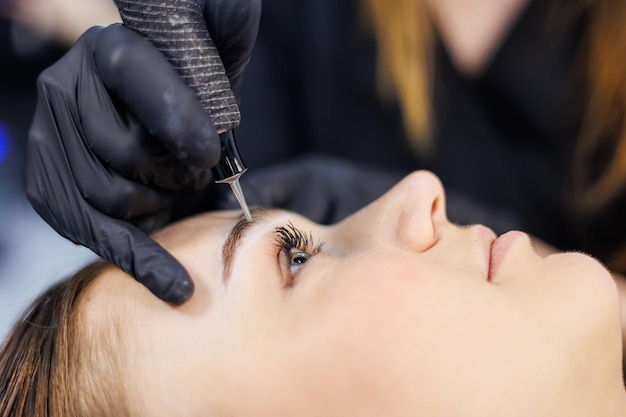 Photo permanent makeup of eyebrows woman professional permanent makeup artist with a permanent makeup tool a young permanent makeup artist makes permanent eyebrow makeup