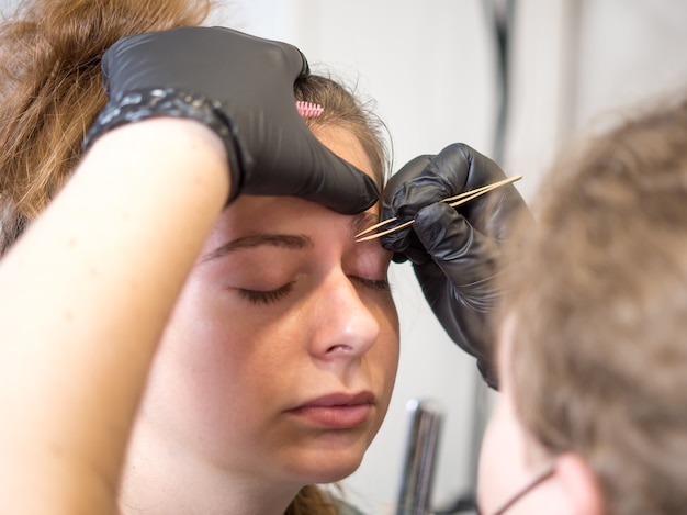 Permanente make-up. Mooie jonge vrouw die de procedure van de wenkbrauwcorrectie ontvangt. De wenkbrauwen van een meisje epileren met een pincet