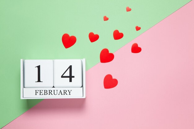 Perpetual calendar with date of February 14, red different-sized confetti hearts on two-color background pink and green. Flat lay. Top view.