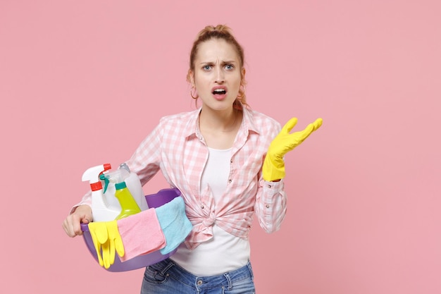 Perplex jonge vrouw huisvrouw in rubberen handschoenen houden bekken met wasmiddelflessen wassen reinigingsmiddelen doen huishoudelijk werk geïsoleerd op roze achtergrond studio portret. Huishoudelijk concept. Handen spreiden.