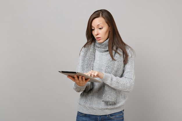 Perplex jonge vrouw in grijze trui, sjaal houden en gebruiken van tablet pc-computer geïsoleerd op een grijze achtergrond. Gezonde levensstijl, online behandelingsadvies, concept voor het koude seizoen. Bespotten kopie ruimte.