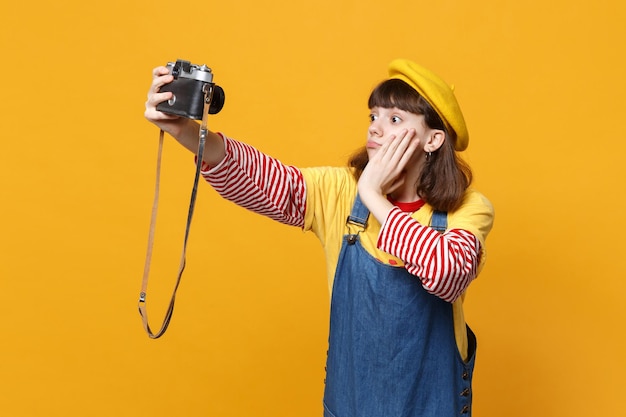 Perplex meisje tiener in Franse baret doen selfie geschoten op retro vintage fotocamera, hand op gezicht geïsoleerd op gele achtergrond. Mensen oprechte emoties, lifestyle concept. Bespotten kopie ruimte.