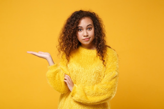 Perplex verwarde jonge afro-amerikaanse meisje in bont trui poseren geïsoleerd op geel oranje achtergrond in studio. mensen oprechte emoties levensstijl concept. bespotten kopie ruimte. wijzende hand opzij.