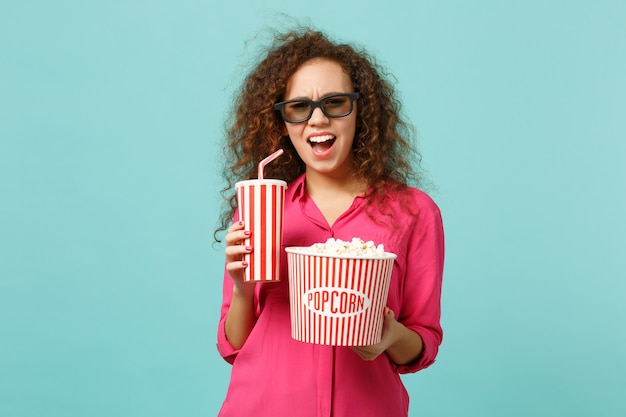 Perplexed african girl in 3d imax glasses watching movie film hold popcorn cup of soda isolated on blue turquoise background in studio. People emotions in cinema lifestyle concept. Mock up copy space.