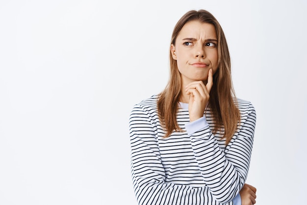 Perplexed blond girl having doubts thinking and looking aside with frowning serious face standing thoughtful and suspicious against white background