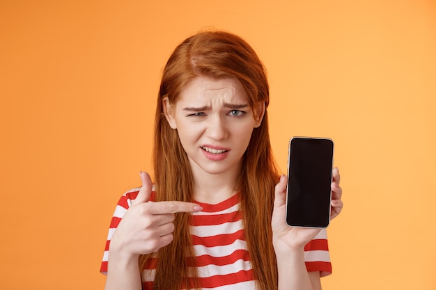 Photo perplexed disappointed redhead girl show smartphone screen app pointing mobile display unsure full d...