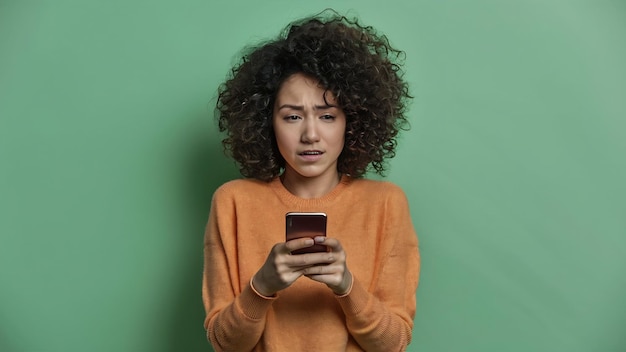 Perplexed hesitant curly haired woman shrugs shoulder being not sure uses smartphone device concent