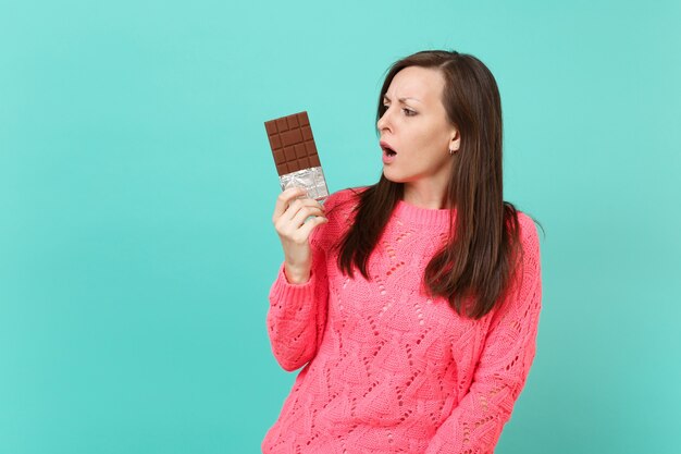 Perplexed puzzled young woman in knitted pink sweater holding in hand looking on chocolate bar isolated on blue turquoise wall background studio portrait. People lifestyle concept. Mock up copy space.