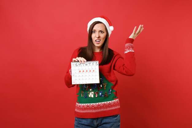 Perplexed Santa girl holding female periods calendar for checking menstruation days isolated on red background. Medical healthcare gynecological. Happy New Year 2019 celebration holiday party concept.