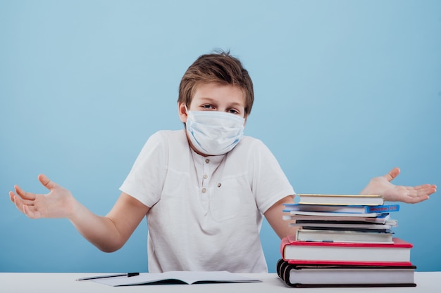 Perplexed schoolboy with medical mask on his face holding notebooks and books in his hand looking at...