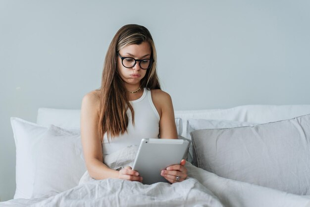 Perplexed young Hispanic woman sitting on bed awakes holds tablet looks at screen puffs out cheeks wears glasses Pensive caucasian girl checks her overloaded agenda Tired student