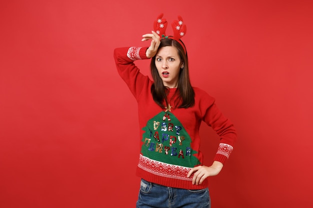 Perplexed young Santa girl in knitted sweater, fun decorative deer horns putting hand on forehead isolated on red background. Happy New Year 2019 celebration holiday party concept. Mock up copy space.