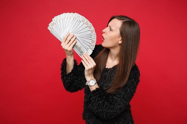 Perplexed young woman in black fur sweater looking on fan of money in dollar banknotes, cash money isolated on red background in studio. People sincere emotions, lifestyle concept. Mock up copy space.