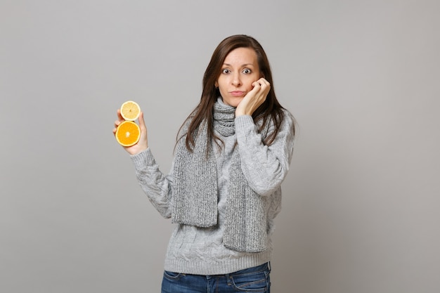 Perplexed young woman in gray sweater, scarf put hand on cheek hold lemon orange isolated on grey background. Healthy fashion lifestyle people sincere emotions cold season concept. Mock up copy space.