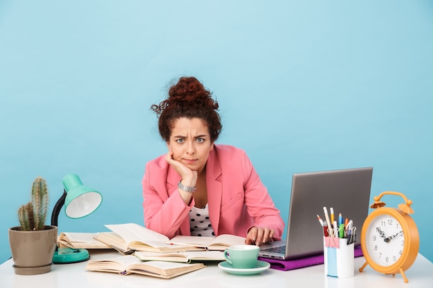 perplexed young woman looking at camera while working at desk isolated over blue wall