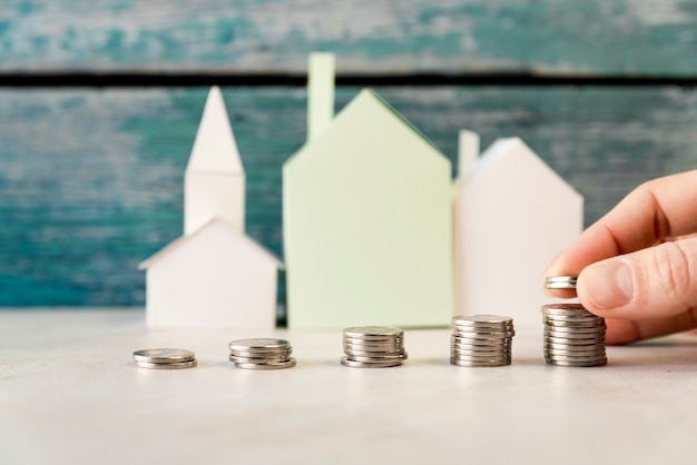 A person arranging the increasing coins in front of paper houses on white surface