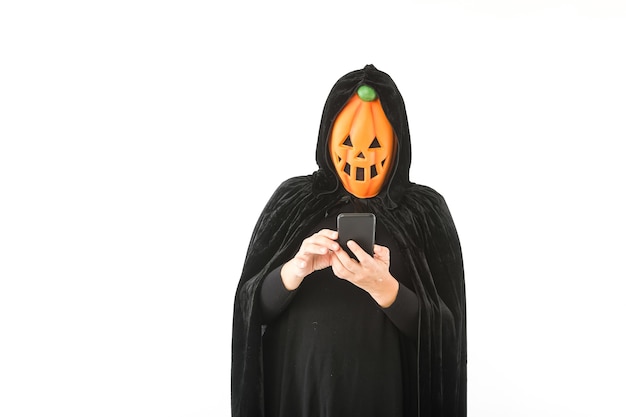 Person dressed in pumpkin Jack-o.-lantern mask and hooded velvet cape, looking at his mobile phone, on white background. Carnival, Halloween and day of the dead concept.