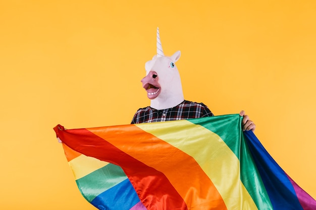 Person dressed in unicorn mask with plaid shirt holding rainbow lgtbq flag on yellow background Concept of gay pride transsexuality and lgtbq rights