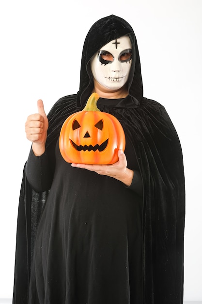 Фото Человек, одетый в маску с крестом на лбу и черный бархатный плащ, держит тыквенный фонарь и поднимает большой палец на белом фоне. карнавал, хэллоуин и день мертвых концепции.