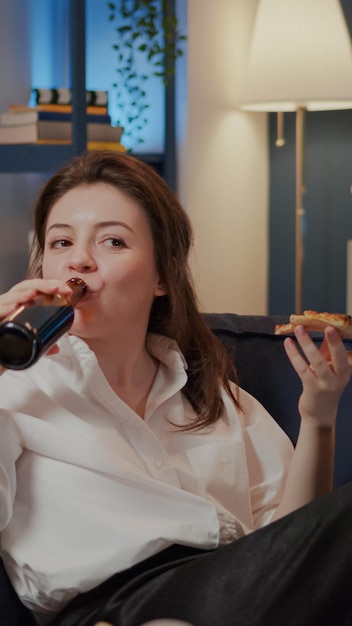 Person eating slice of pizza while watching comedy on tv and\
laughing on couch at home. caucasian woman having fun with movie on\
television, enjoying fast food takeaway and bottle of beer